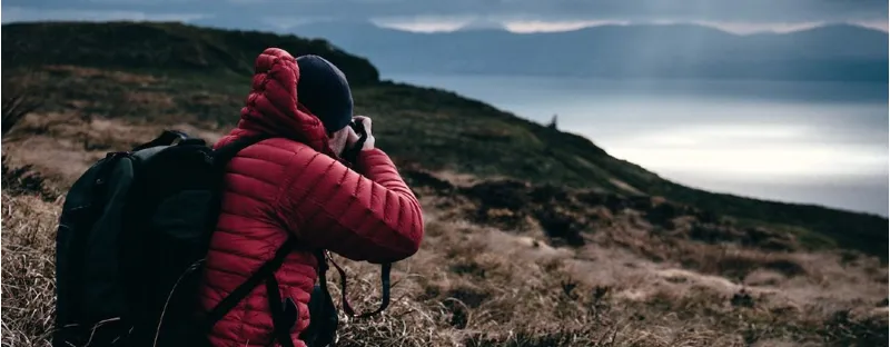 Man wearing padded jacket on hillside taking a photo of mountains